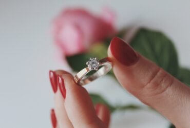 A closeup shot of a female holding a gold diamond ring with a pink rose in a blurred background